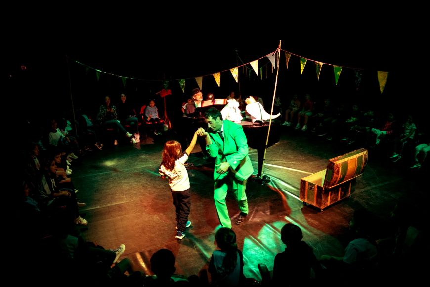 L'actor Pep Farrés està ballant al mig de l'escenari amb un infant que ha sortit voluntari del públic. El músic Guillem Martí toca el piano de cua.