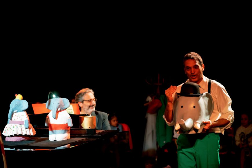 L'actor Pep Farrés té a les mans un cap d'elefant i està parlant. El músic Guillem Martí està tocant el piano tot mirant cap a l'esquerra. Damunt del piano hi ha dues figuretes d'elefants.