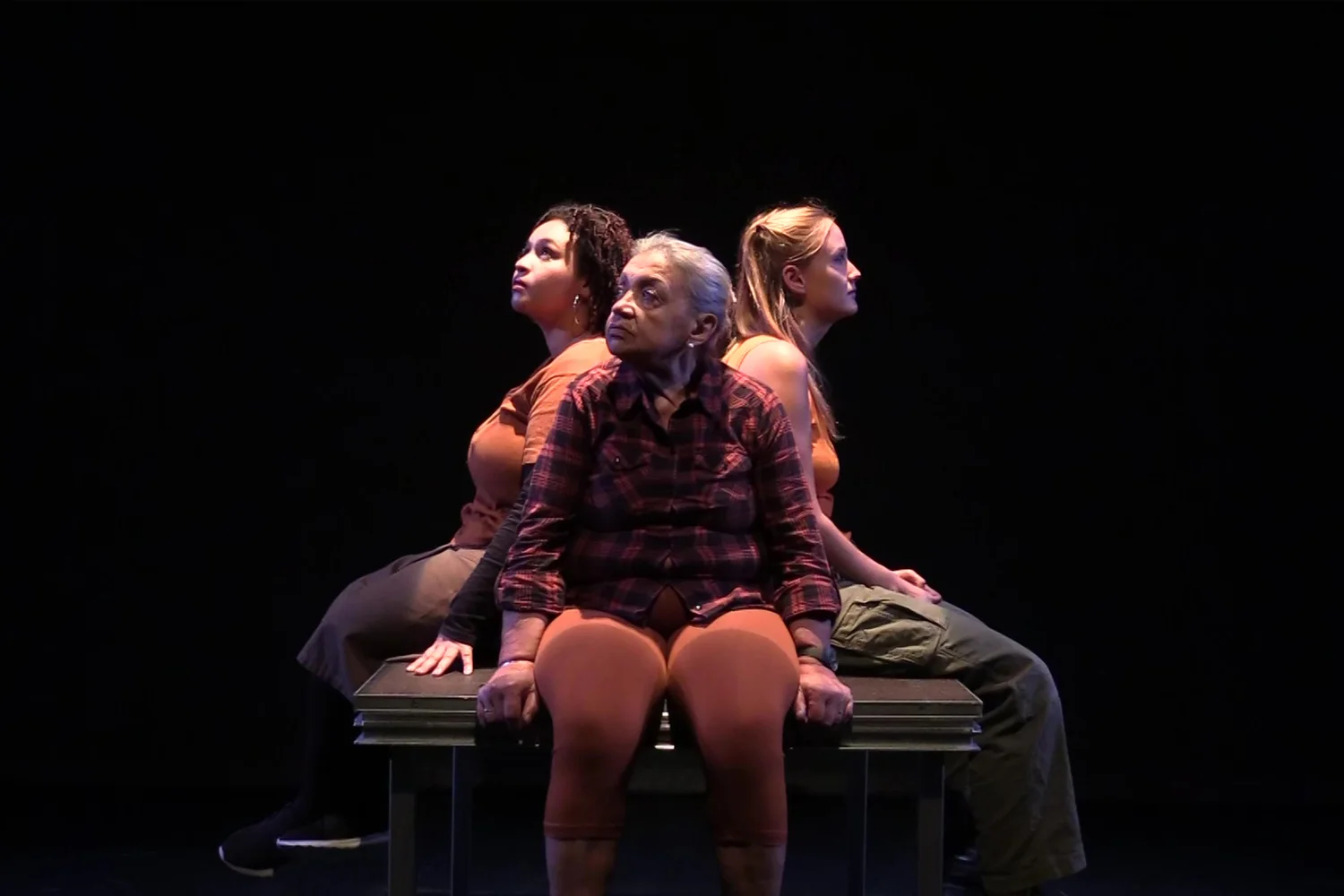 Tres dones assegudes donant-se l'esquena, una de gran, una de mitjana edat i una de jove, il·luminades només elles però envoltades de foscor