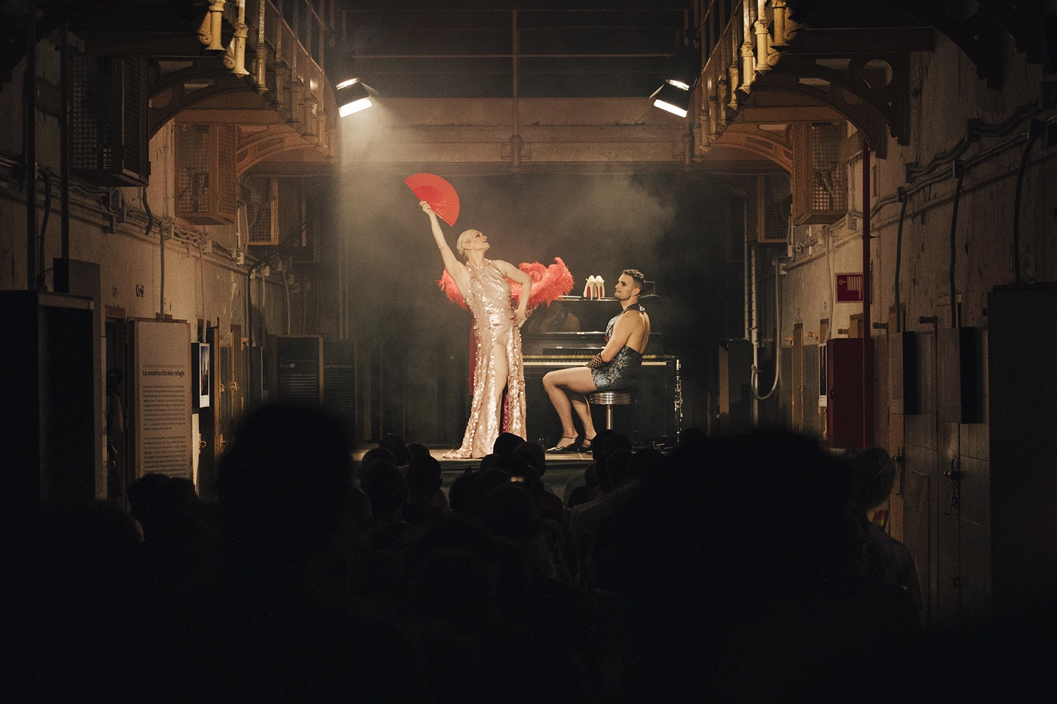 Imatge d'escena on un travesti amb un vestit platejat aixeca un vano vermell al costat d'un piano de paret on hi ha assegut un pianista que se'l mira