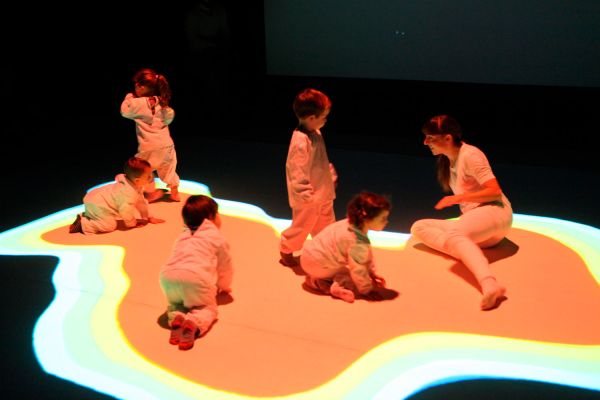 Infants de 2 anys vestits amb una granota blanca i una ballarina també vestida de blanc, enmig d'un escenari negre amb el terra amb una taca de color projectada