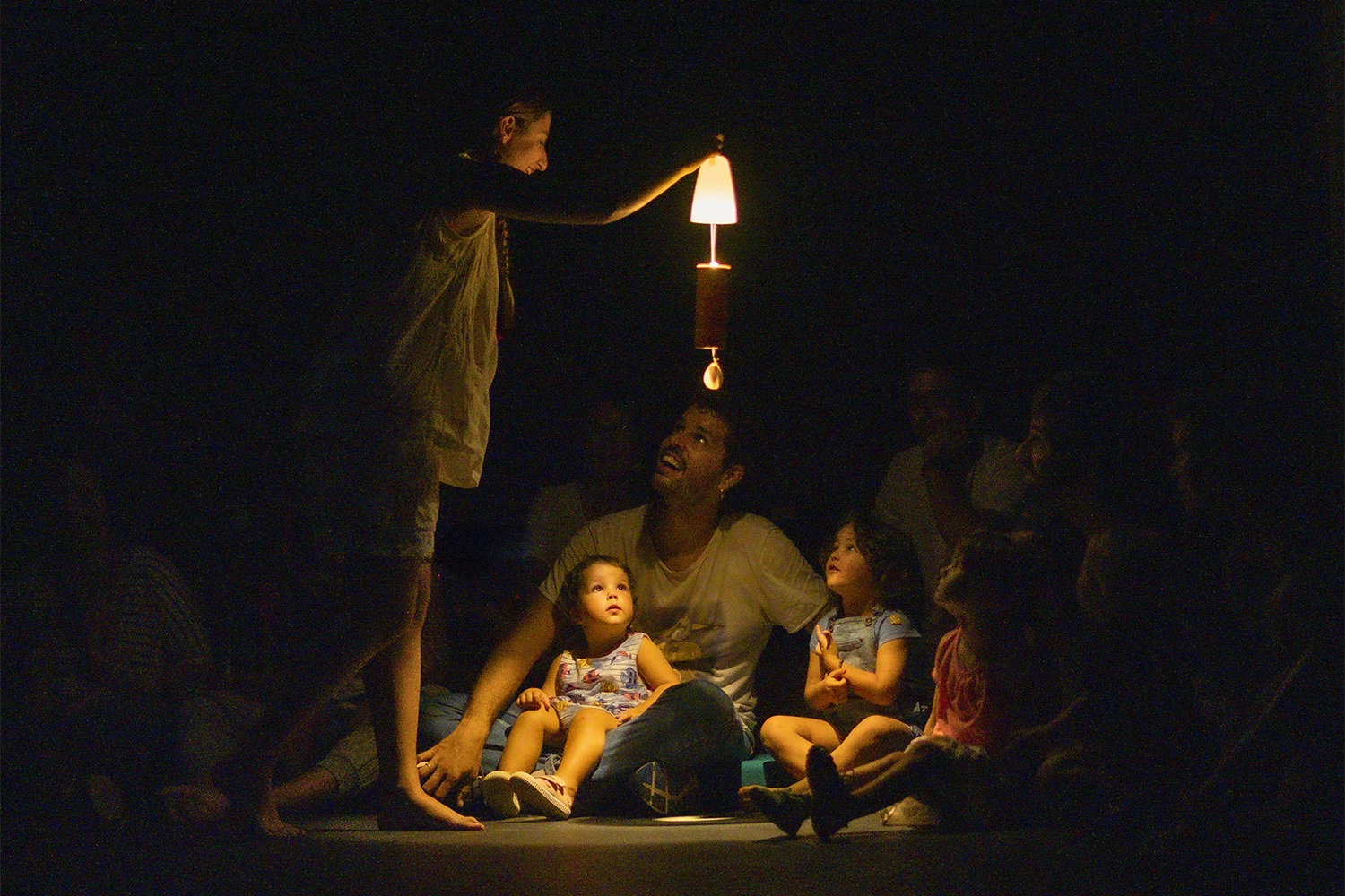 Una família asseguda a terra miren una làmpada encesa que està  per damunt dels seus caps i que sostè una actriu