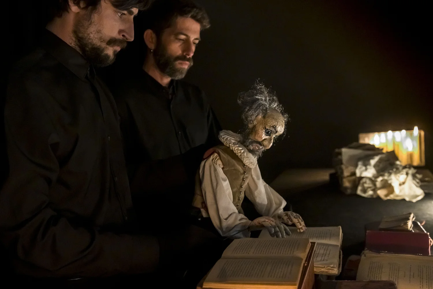 Dos actors vestits de negre observen un titella del Quixto que està llegin llibres a la llum de les espelmes